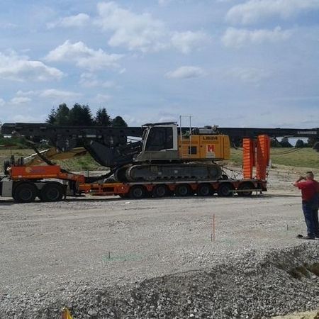 Ein Tieflader, auf dem eine große, grau- und orangefarbene Raupenbagger mit langem Ausleger transportiert wird