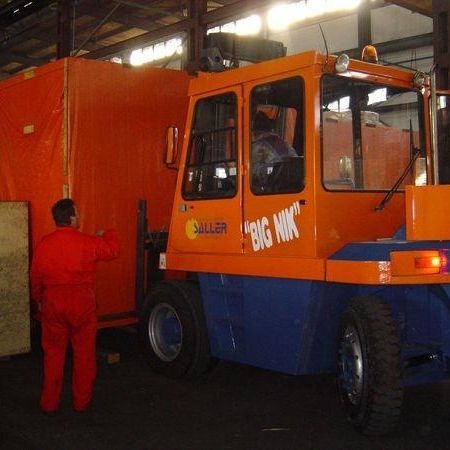 Ein Lagerarbeiter in einem roten Overall bedient einen großen blauen und orangefarbenen Gabelstapler mit der Aufschrift „BIG MK“