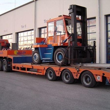 Ein orangefarbener und blauer Gabelstapler ist auf einen Tieflader geladen, der in der Nähe großer Lagertore geparkt ist. Ein zweiter Lkw ist in der Nähe zu sehen
