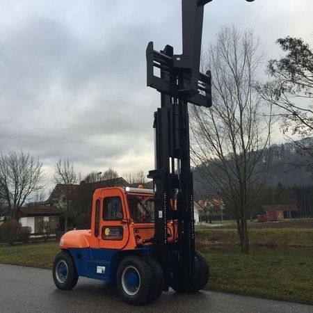 Ein orangefarbener und blauer Gabelstapler steht auf einer wolkenverhangenen Straße und präsentiert seinen langen Hebearm vor einer Kulisse aus Bäumen und Gebäuden