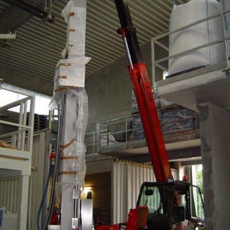 Ein roter Teleskoplader hebt einen hohen, verpackten Gegenstand in einem Lagerhaus an, das von Maschinen und Lagerbereichen umgeben ist.