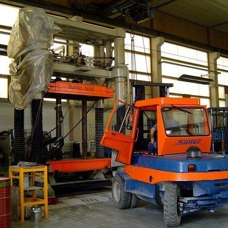 Ein leuchtend orangefarbener Gabelstapler steht neben einer großen, teilweise abgedeckten Maschine in einer geräumigen Werkstatt